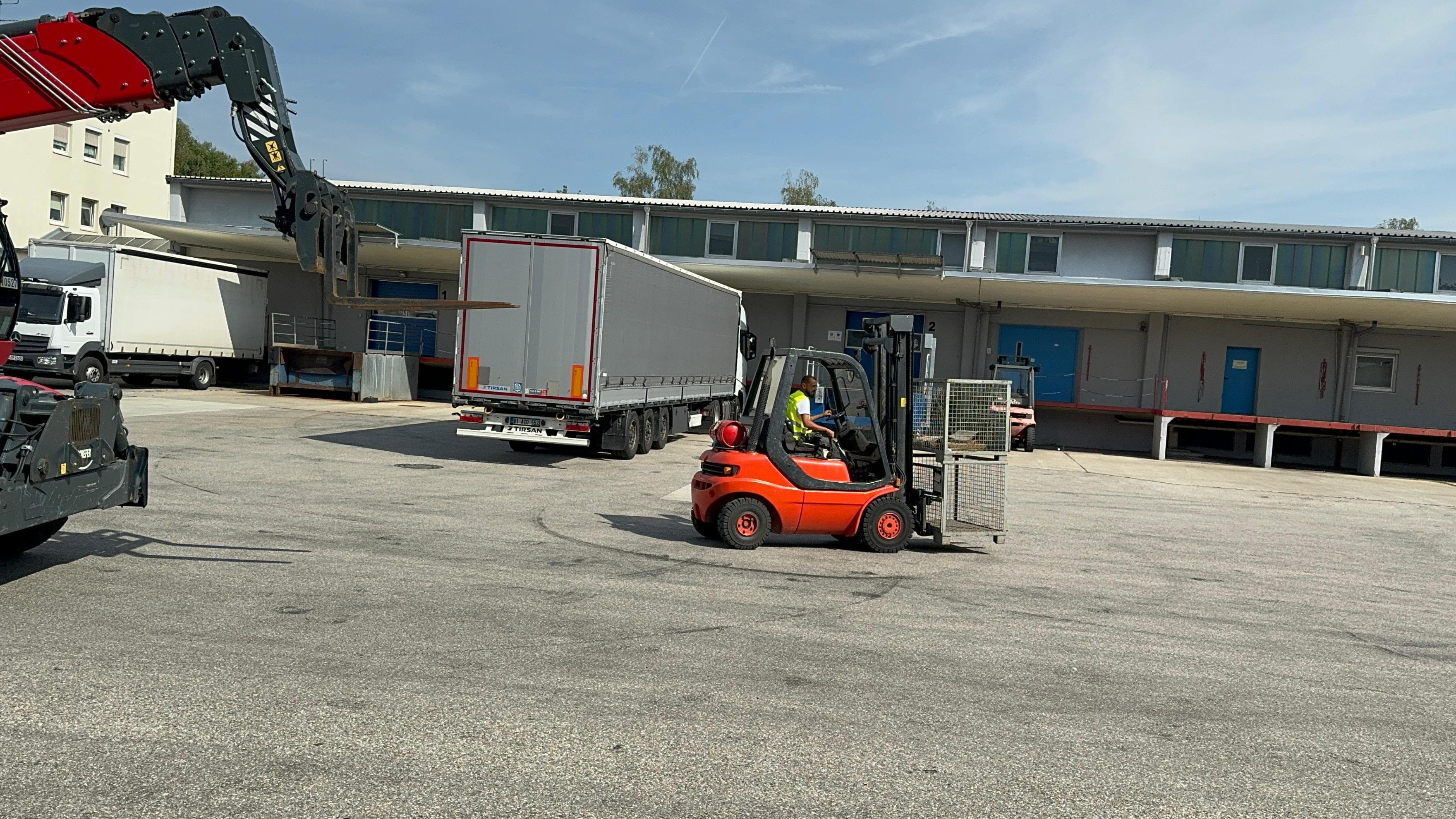 Ein roter Gabelstapler und ein Teleskopstapler arbeiten gemeinsam auf einem Lagerhof, ein Lkw im Hintergrund.